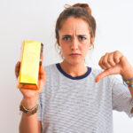 Young beautiful redhead woman holding gold ingot over isolated background with angry face, negative sign showing dislike with thumbs down, rejection concept